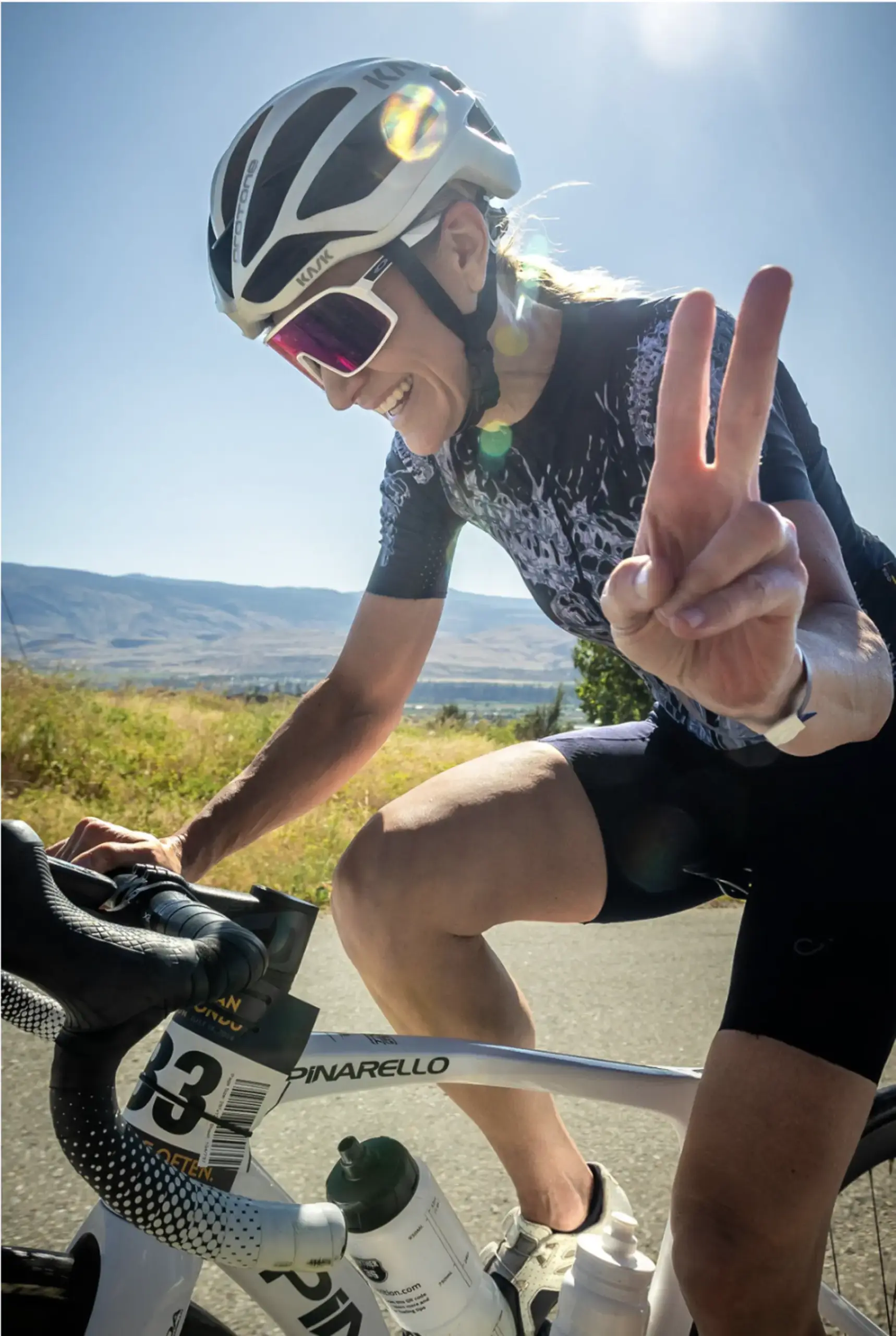 A cyclist wearing a helmet and sunglasses smiles and flashes a peace sign while riding a road bike on a sunny day, with hills and blue sky in the background.