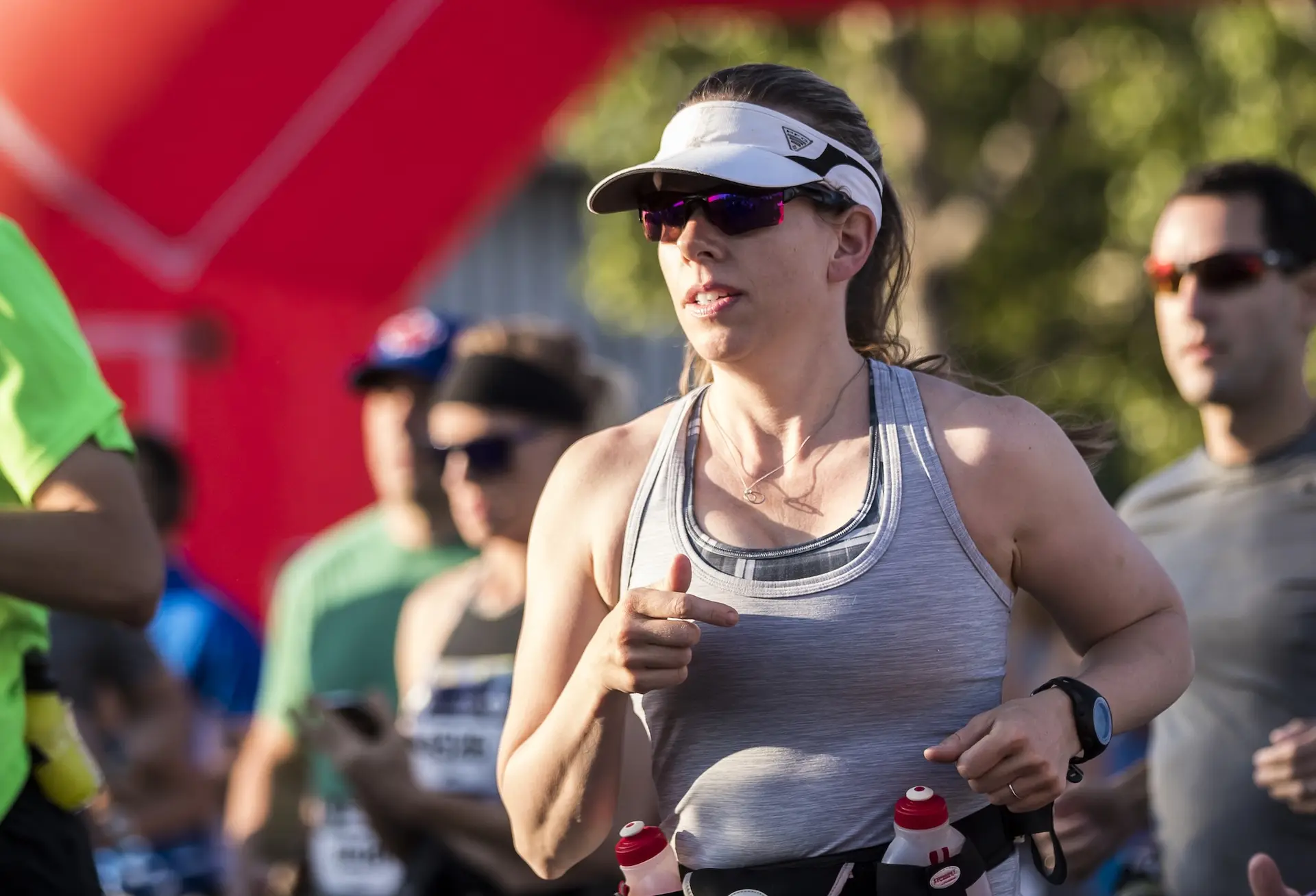 A woman wearing sunglasses, a visor, and a gray tank top runs in a race, holding water bottles at her waist. Other runners are visible in the background, outdoors in daylight.