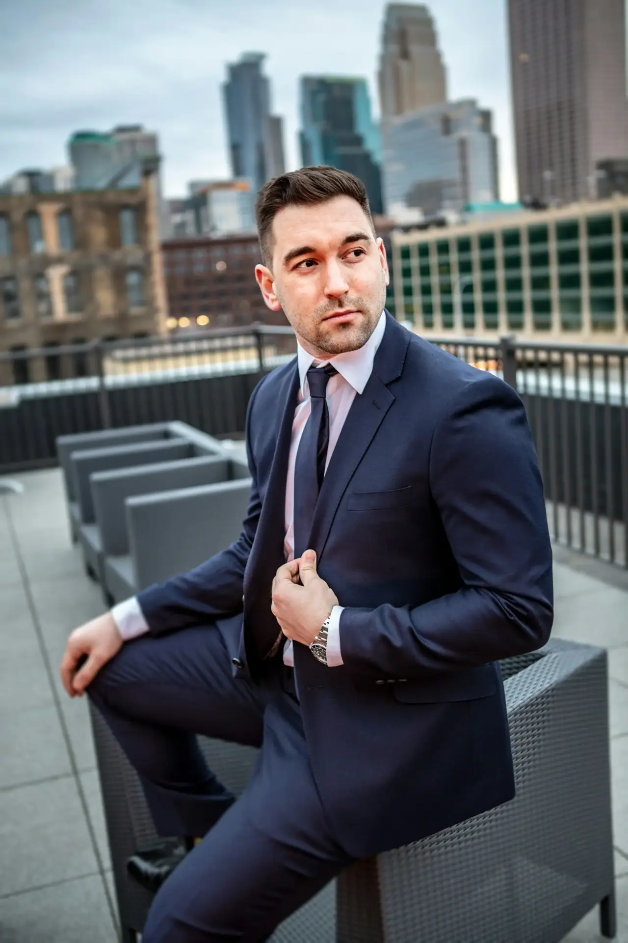 A man in a navy blue suit and tie sits on a gray outdoor chair on a rooftop patio with city buildings in the background. He is looking to the side with one hand holding his suit jacket.