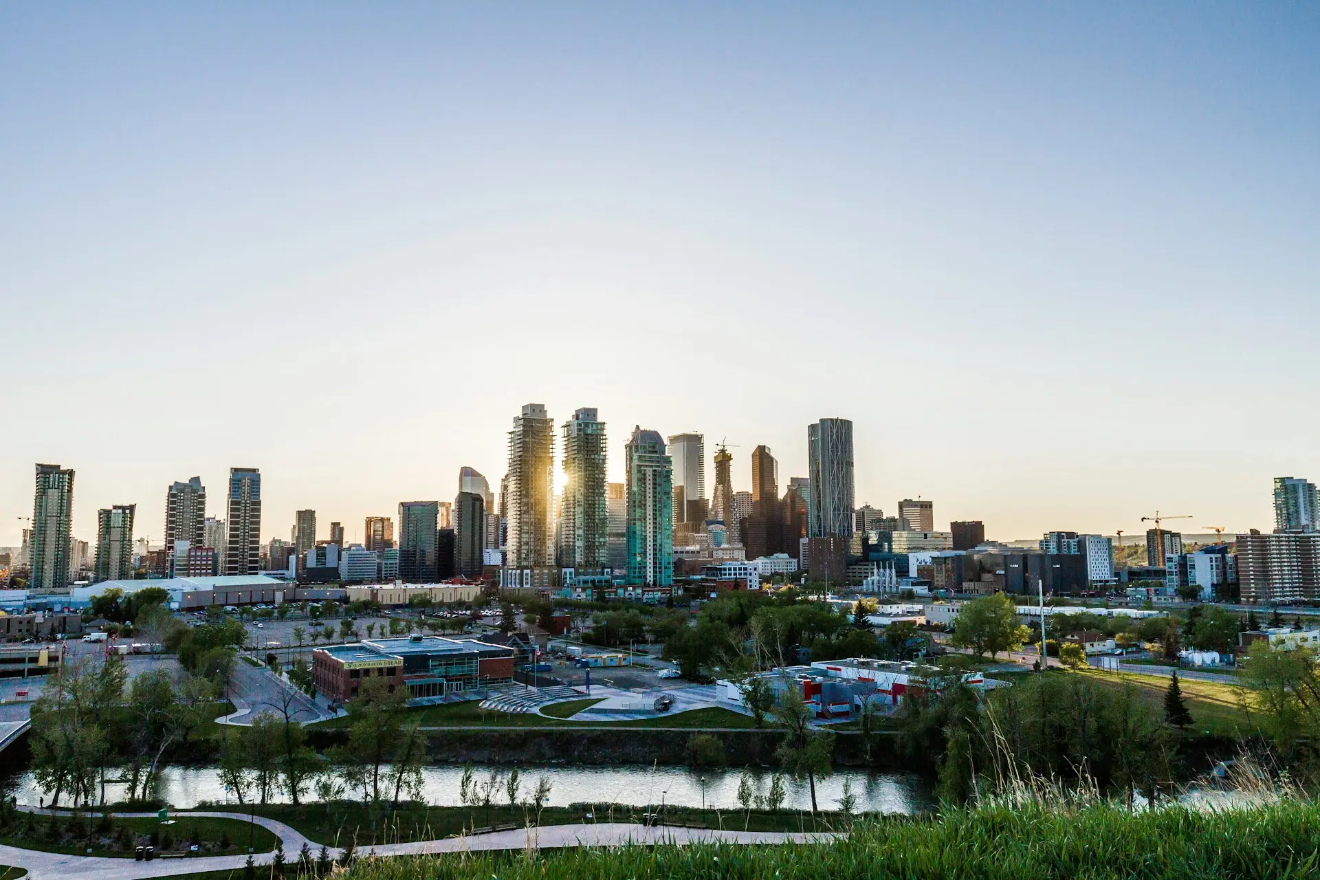 A city skyline with tall modern buildings and a river in the foreground under a clear blue sky, photographed at sunrise or sunset. Grass and trees are visible near the water.