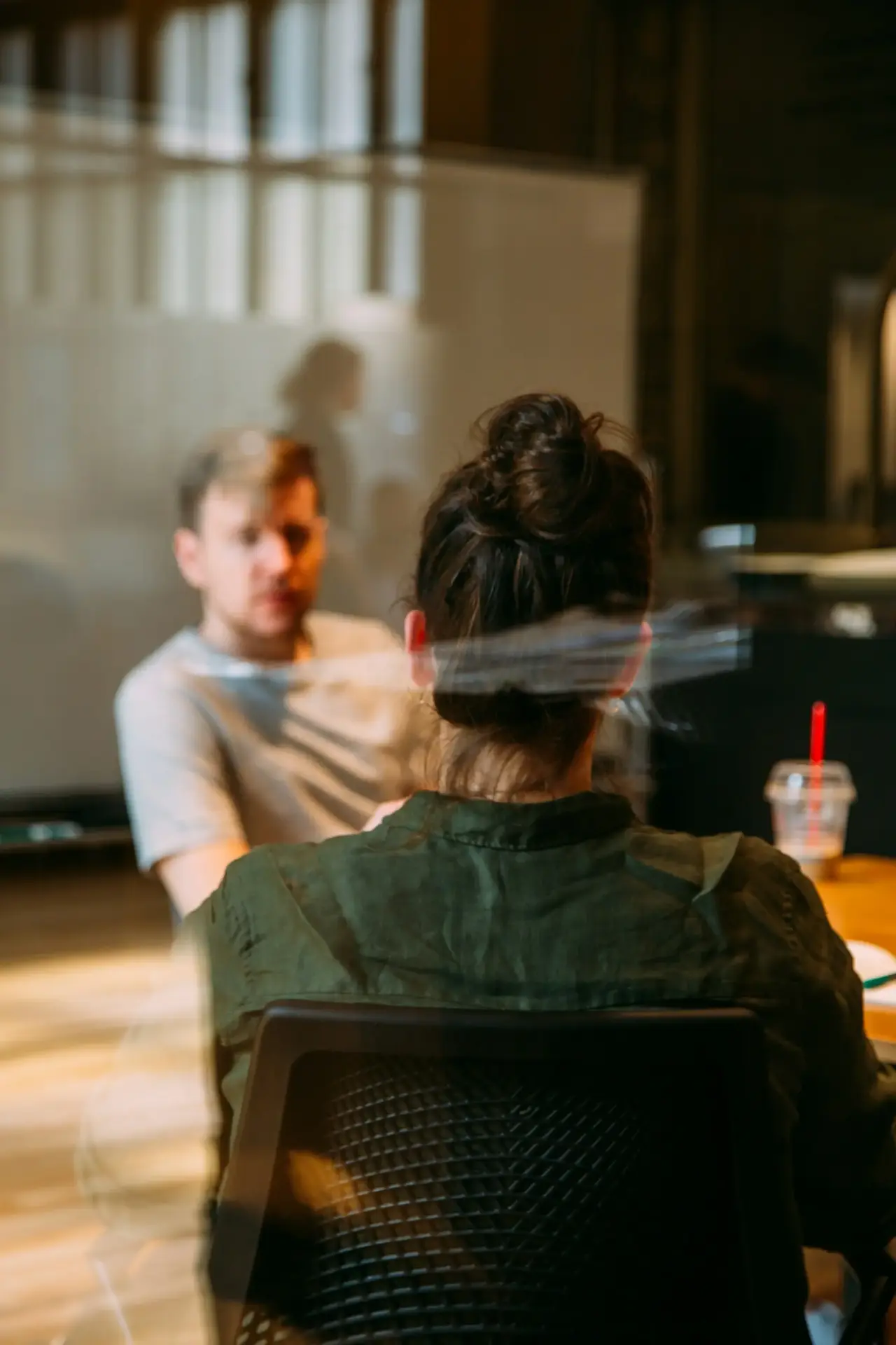 A man and a woman sit across from each other at a table in an office setting. The man faces the camera while the woman is seen from behind. There is a drink with a red straw on the table.